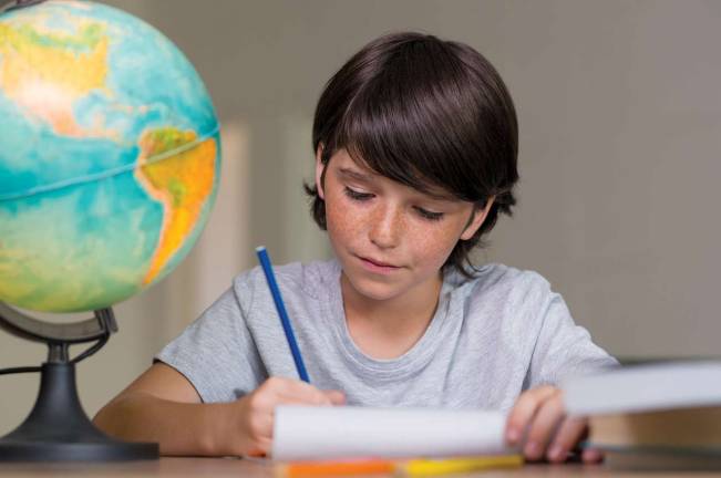 Closeup of little boy doing homework beside globe of earth. Portrait of concentrated boy studying geography. Cute boy doing his homework at desk.