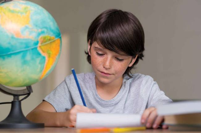 Location, location, location Closeup of little boy doing homework beside globe of earth. Portrait of concentrated boy studying geography. Cute boy doing his homework at desk.