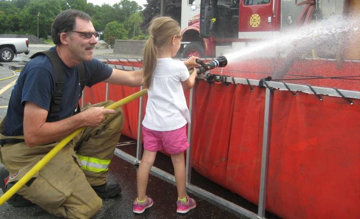 PHOTO BY JANET REDYKE Six-year-old Erika, with the help of fireman Ben Cosh of the Wantage Beemerville Company, has a blast working the fire hose.